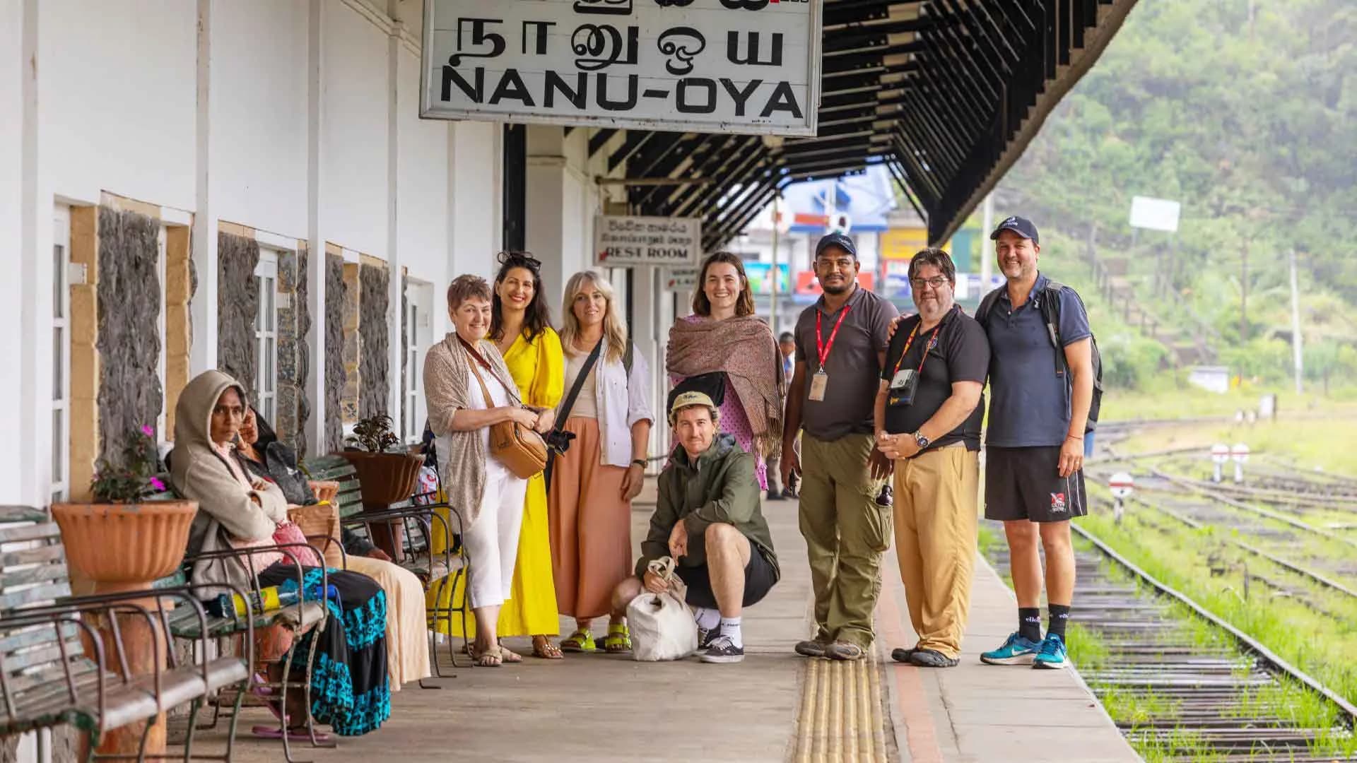 A group of travelers and guides posing together at a historic railway station platform, featuring a train car in the background.