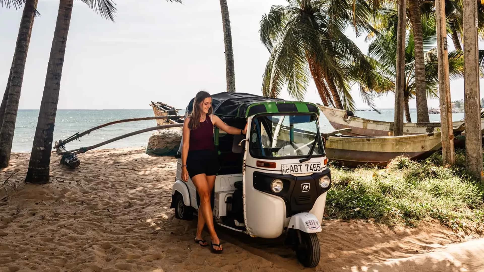 A woman with a backpack standing next to a black tuk-tuk taxi parked on a tropical beach near palm trees.