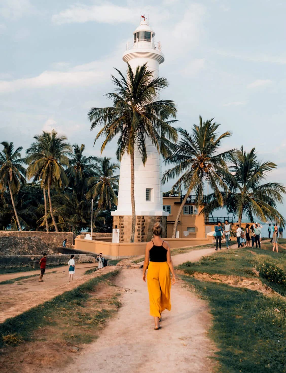 A traveler in a yellow outfit seen from behind, walking toward a white, historic lighthouse near palm trees at twilight.